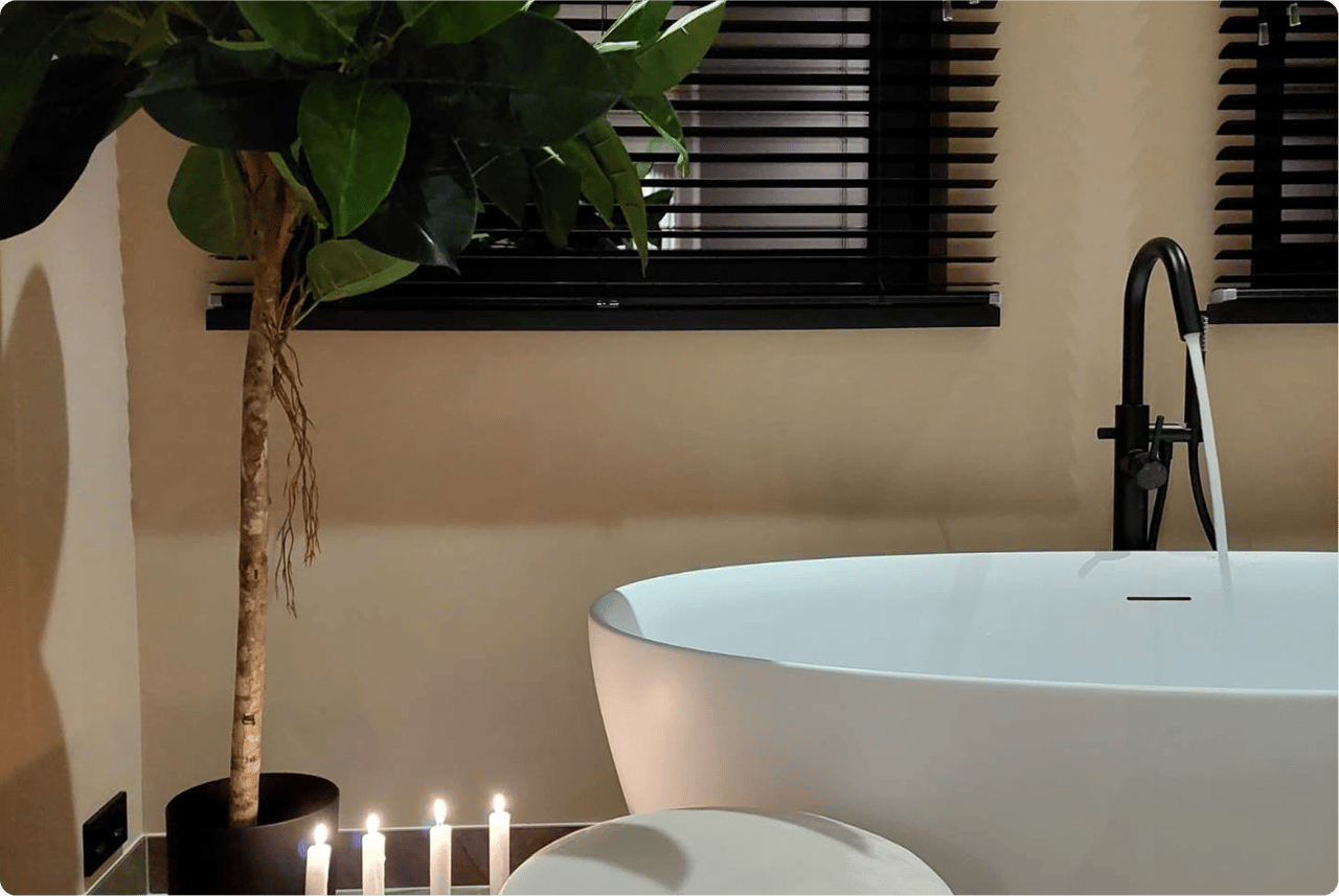 Stimmungsvolles x²o Bad mit freistehender weißer Badewanne, schwarzer Standarmatur und sanfter Beleuchtung. Sechs brennende Kerzen und eine große grüne Zimmerpflanze sorgen für eine beruhigende Atmosphäre. Im Hintergrund sind schwarze Jalousien vor dem Fenster zu sehen.